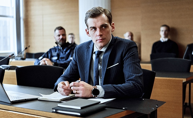 Näyttelijä Lauri Tilkanen istuu Pohjolan laki -sarjan kuvauksissa oikeussalissa. Pöydällä hänen edessään on muistivihkoja ja tietokone.